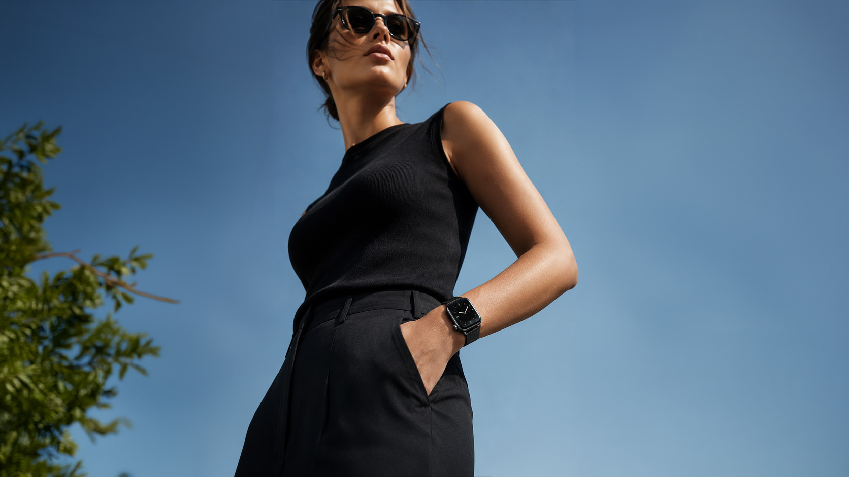 A woman in a black sleeveless dress and sunglasses, shot from a low angle against a clear blue sky.